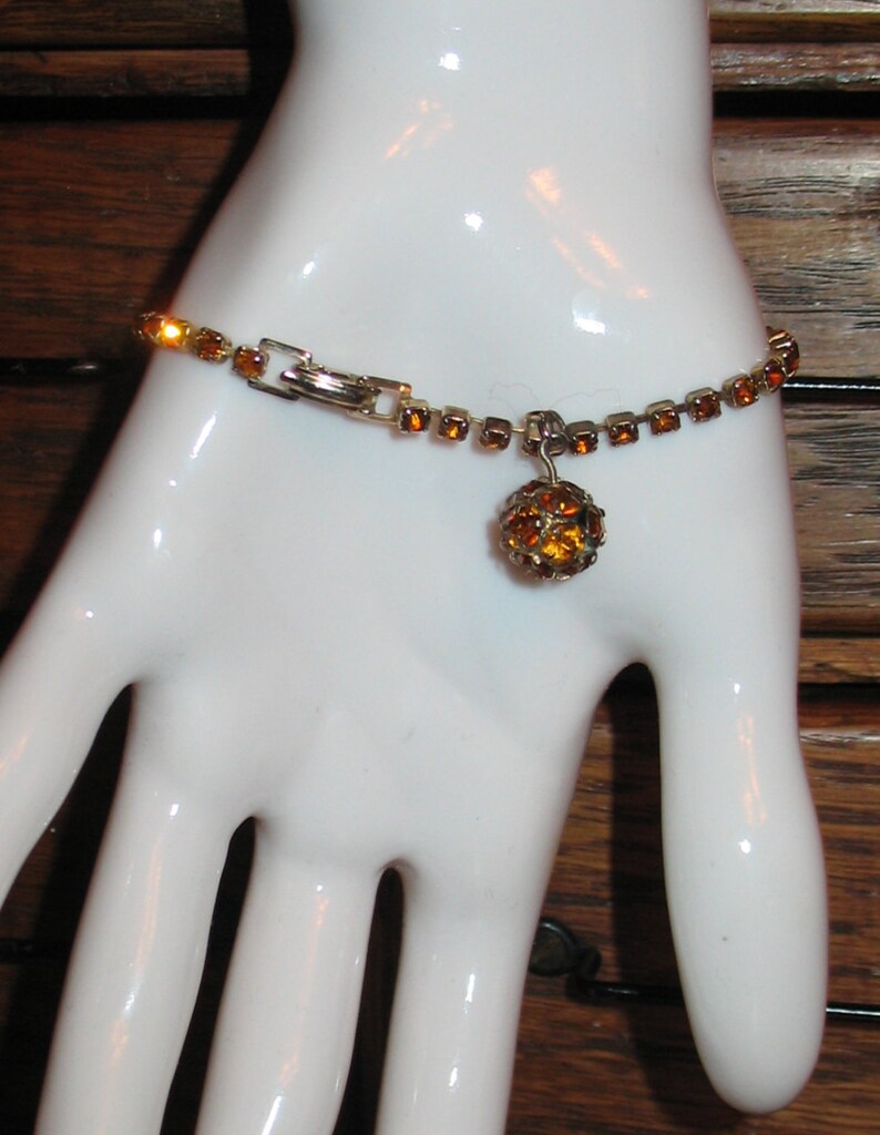 May include: A gold tone bracelet with a dangling amber colored rhinestone ball charm. The bracelet is made of small square rhinestones.