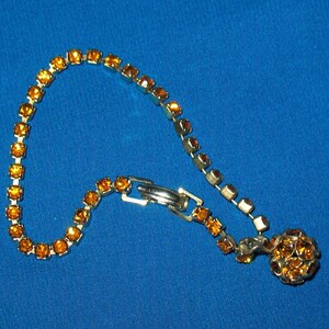 May include: A gold tone bracelet with a cluster of amber-colored rhinestones at the end. The bracelet is made of a chain of small, square-shaped rhinestones.