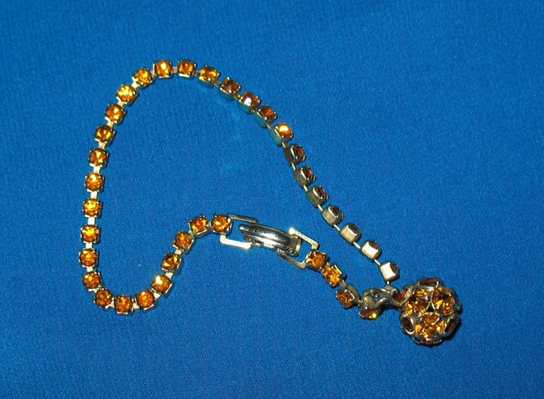 May include: A gold tone bracelet with a cluster of amber-colored rhinestones at the end. The bracelet is made of a chain of small, square-shaped rhinestones.