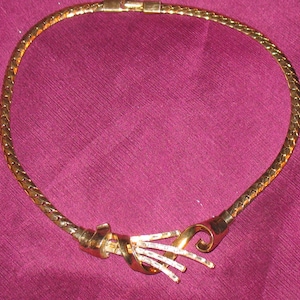 May include: A gold-tone chain necklace with a twisted, ornate clasp. The necklace is made of a braided chain and has a simple, elegant design.