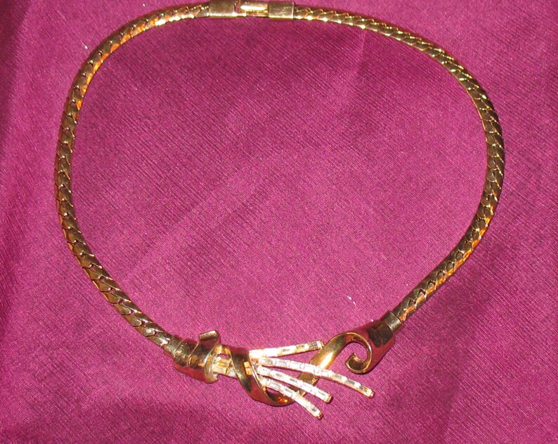 May include: A gold-tone chain necklace with a twisted, ornate clasp. The necklace is made of a braided chain and has a simple, elegant design.