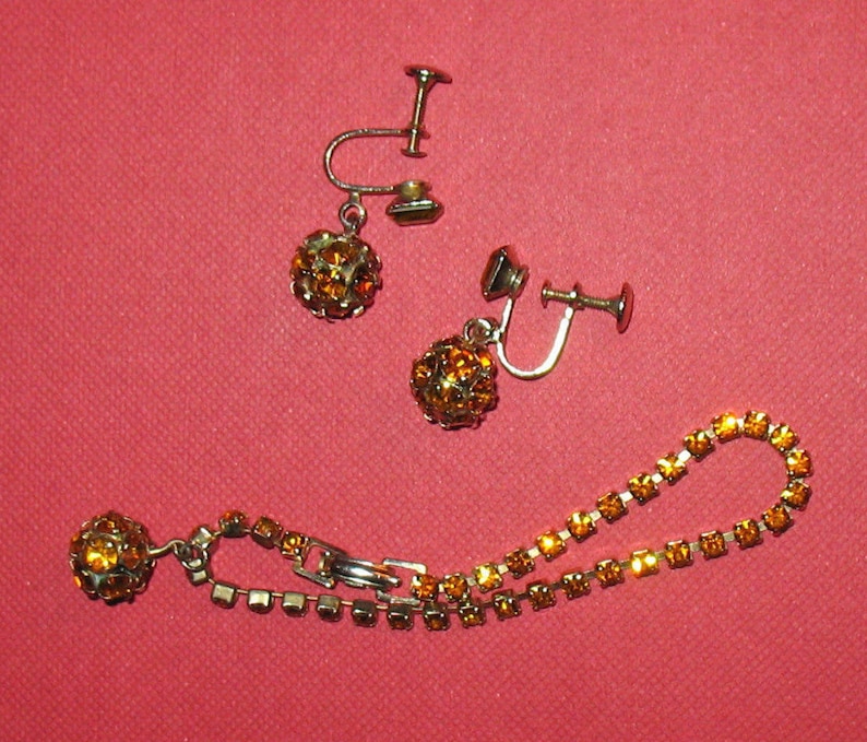 May include: A set of amber rhinestone jewelry including a pair of clip-on earrings and a necklace.