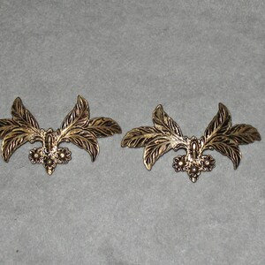 May include: Two gold-toned metal leaf-shaped embellishments with a decorative floral design. Each embellishment has three leaves and a cluster of three small flowers in the center.