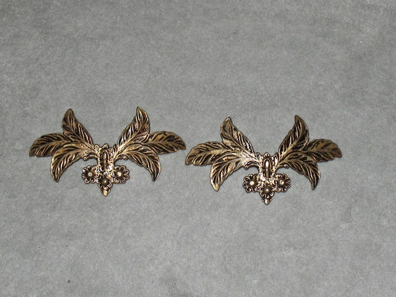 May include: Two gold-toned metal leaf-shaped embellishments with a decorative floral design. Each embellishment has three leaves and a cluster of three small flowers in the center.