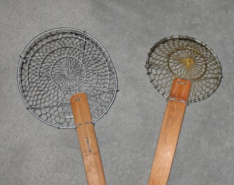 May include: Two round wire mesh kitchen strainers with wooden handles. The strainer on the left is silver and the strainer on the right is gold.
