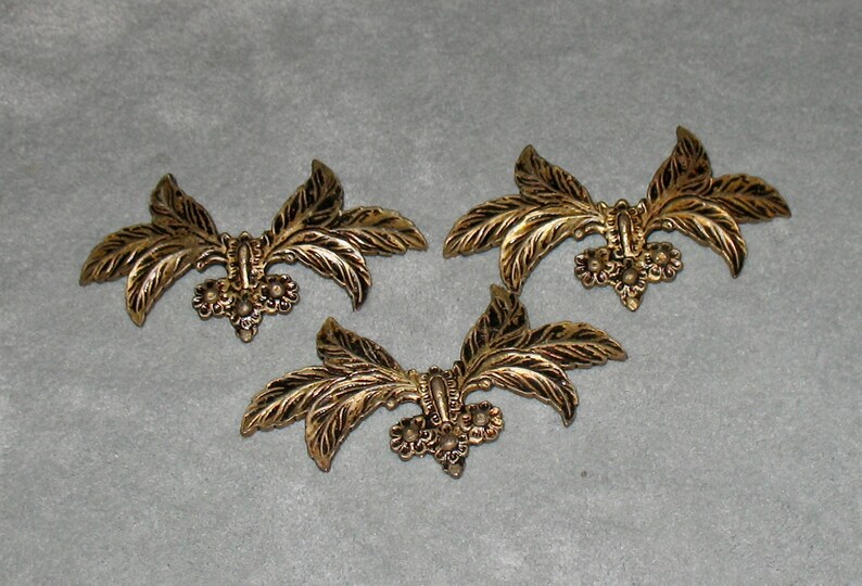 May include: Three gold-toned metal decorative embellishments shaped like stylized leaves with a floral design in the center.