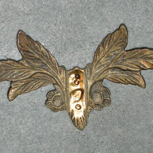 May include: A gold-colored metal decorative piece with a leaf design. The piece has a central section with a raised design and two leaves extending out on either side.