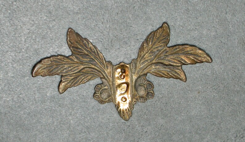 May include: A gold-colored metal decorative piece with a leaf design. The piece has a central section with a raised design and two leaves extending out on either side.