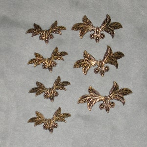 May include: Eight gold-colored metal leaf-shaped embellishments with a decorative center. The embellishments are arranged in rows on a gray surface.