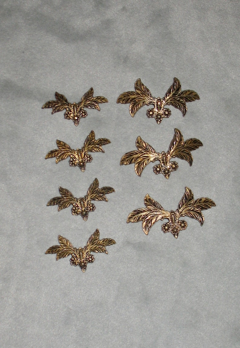 May include: Eight gold-colored metal leaf-shaped embellishments with a decorative center. The embellishments are arranged in rows on a gray surface.
