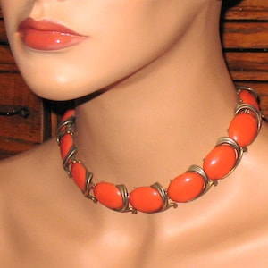 May include: A close-up of a coral-colored necklace with oval-shaped stones set in gold-toned metal. The necklace is worn by a mannequin with a smooth, light-colored skin tone.