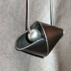 Puede incluir: Un collar con un colgante de plata de dise&ntilde;o geom&eacute;trico negro y cobre. El colgante presenta una bola redonda de plata suspendida dentro de la forma geom&eacute;trica.