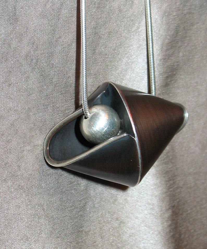 Puede incluir: Un collar con un colgante de plata de dise&ntilde;o geom&eacute;trico negro y cobre. El colgante presenta una bola redonda de plata suspendida dentro de la forma geom&eacute;trica.
