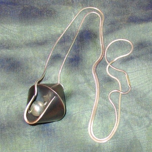 Puede incluir: Un collar de cadena de plata con un colgante de metal gris oscuro. El colgante tiene forma de tri&aacute;ngulo plegado y tiene una peque&ntilde;a bola de plata en su interior.