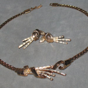 May include: A gold tone necklace and matching earrings set. The necklace is a chain with a decorative knot and a cluster of gold tone spikes. The earrings are a similar design with a knot and spikes.