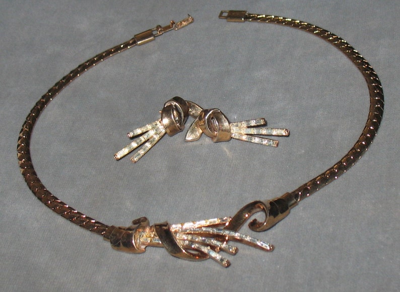 May include: A gold tone necklace and matching earrings set. The necklace is a chain with a decorative knot and a cluster of gold tone spikes. The earrings are a similar design with a knot and spikes.