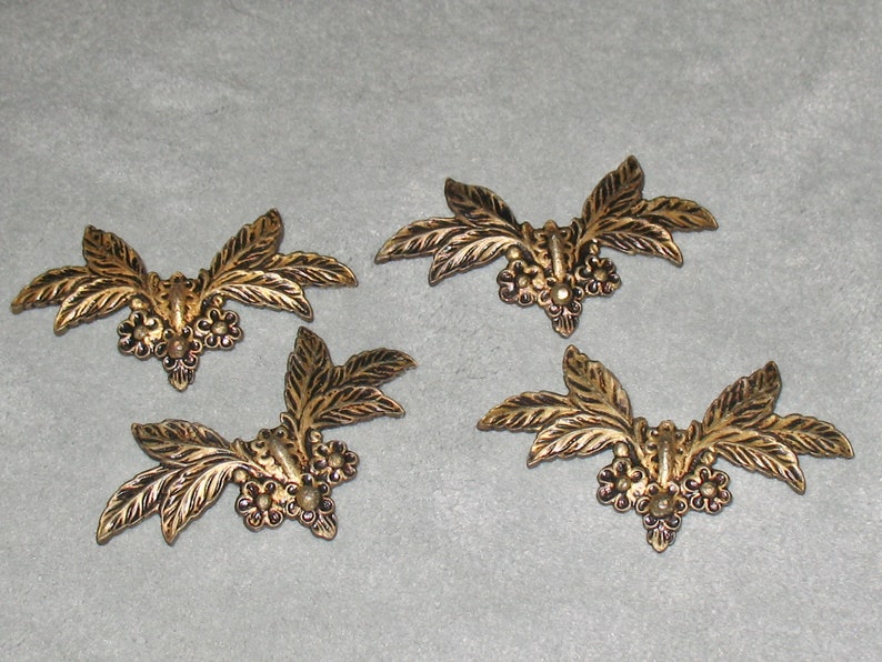 May include: Four gold-colored metal leaf and flower embellishments. Each embellishment features a central cluster of three small flowers with a raised center, surrounded by two sets of three leaves.