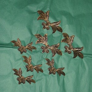 May include: Eight gold-colored metal leaf-shaped embellishments with small flower details. The embellishments are arranged in a triangular pattern on a green background.