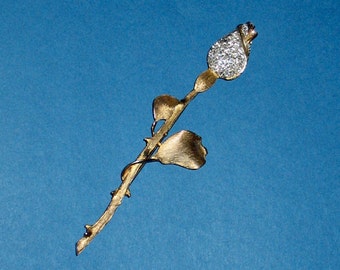 Puede incluir: Un broche de rosa dorado con un capullo incrustado de diamantes de imitación y un tallo y hojas estilizados.