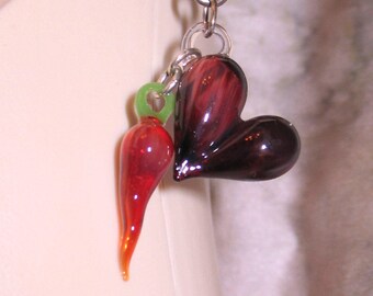 May include: A red glass heart charm with a red glass chili pepper charm attached to it. The charms are hanging from a silver chain.