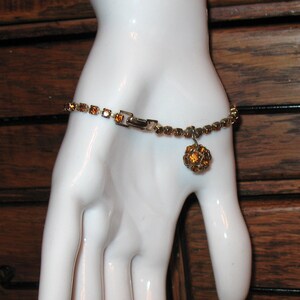 May include: A gold tone bracelet with a dangling amber rhinestone ball charm. The bracelet is made of small, square rhinestones.