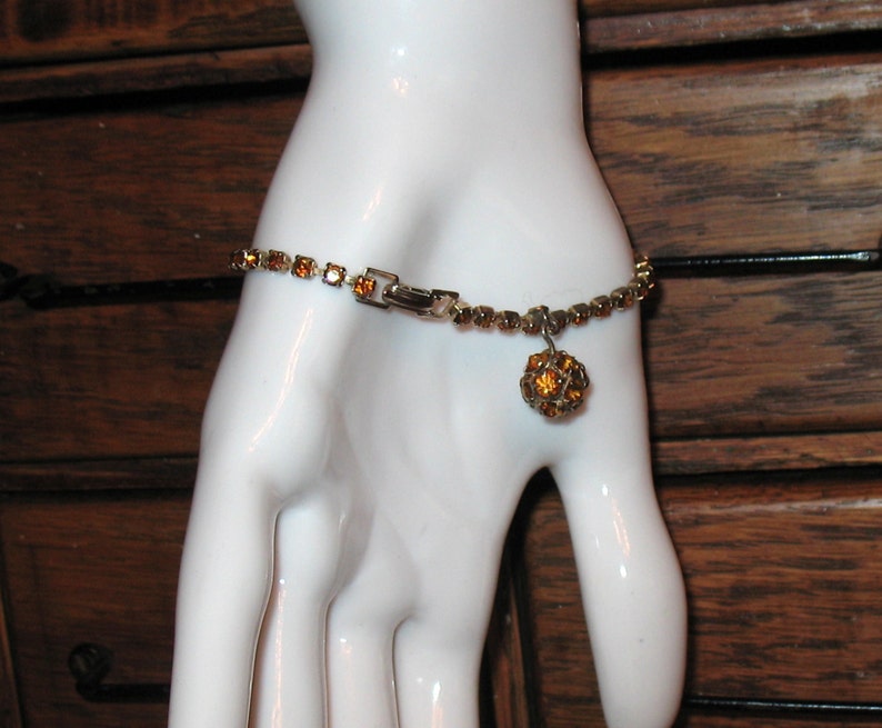 May include: A gold tone bracelet with a dangling amber rhinestone ball charm. The bracelet is made of small, square rhinestones.