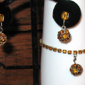 May include: A set of amber rhinestone jewelry featuring a bracelet with a dangling ball charm and matching earrings with a single rectangular stone and a dangling ball charm.