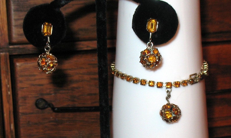 May include: A set of amber rhinestone jewelry featuring a bracelet with a dangling ball charm and matching earrings with a single rectangular stone and a dangling ball charm.