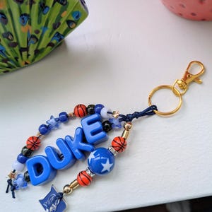 May include: A blue and gold keychain with the word "DUKE" spelled out in blue block letters. The keychain features basketball-themed beads, star-shaped beads, and a gold clasp. A small blue and white emblem hangs from the bottom.