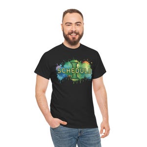May include: Black t-shirt with the word "SCHEDULE" in a green, watercolor-style font, with a large green "I" in the center. The text is set against a colorful splash of blue, green, and yellow paint.