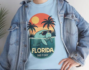 May include: Light blue t-shirt featuring a cartoon manatee wearing sunglasses, set against a sunset with palm trees. The text "FLORIDA TAKE IT EASY" is printed below the manatee. The shirt is made of a soft material.