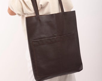 May include: A brown leather tote bag with a large front pocket and a brown leather strap. The bag is being held by a person wearing a white sweater.