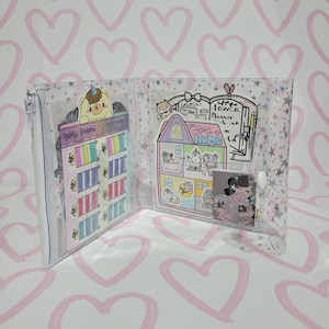 May include: A clear plastic binder with a zipper closure. The binder has a star and glitter pattern. The inside of the binder has a variety of colorful stickers and a page with the text "Stay Home" and "I am a Planner" written on it.