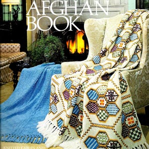 May include: The cover of "The Afghan Book" shows a crocheted afghan draped over a chair. The afghan has a white base with colourful geometric patterns and a fringed edge. A blue knitted blanket is also visible. The book title is prominently displayed.