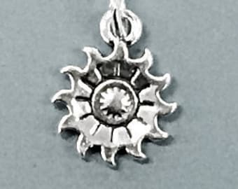 May include: Silver charm in the shape of a sun with a detailed design. The sun has a center with a small circle and rays extending outward.