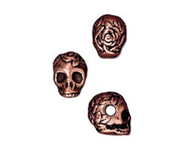 May include: Three copper-colored skull beads with floral designs. The skulls are detailed with eye sockets and a rose design.