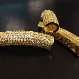 May include: Three gold-colored, curved jewelry components. Each piece is encrusted with small, clear gemstones. The components have a hollow, open design on the inside. The background is a dark, reflective surface.