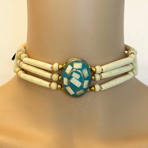 May include: A white bone bead choker necklace with a turquoise and gold oval pendant. The pendant has a geometric design with white and gold accents.