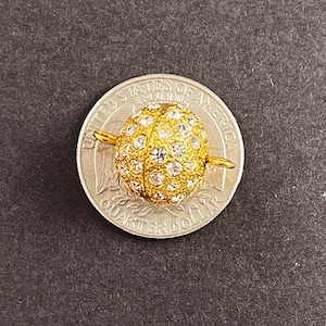 Op de afbeelding: Een gouden metalen sluiting met een rond, met heldere kristallen bezet ontwerp. De sluiting wordt getoond naast een Amerikaanse kwart dollar munt voor groottevergelijking.