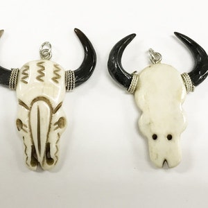 Peut inclure: Deux pendentifs en os de cr&acirc;ne blanc avec des accents de corne noire. Les pendentifs ont des boucles argent&eacute;es pour accrocher. Un cr&acirc;ne a un motif sculpt&eacute; sur le devant.