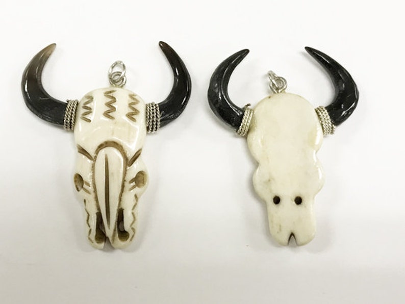 Peut inclure: Deux pendentifs en os de cr&acirc;ne blanc avec des accents de corne noire. Les pendentifs ont des boucles argent&eacute;es pour accrocher. Un cr&acirc;ne a un motif sculpt&eacute; sur le devant.