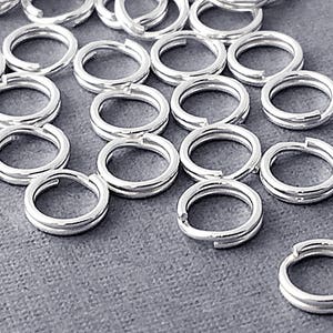 May include: A pile of silver jump rings, a type of jewelry finding, scattered on a gray surface. The rings are all the same size and are made of a shiny metal.