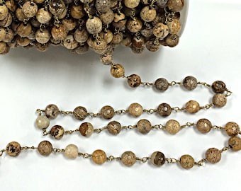 Op de afbeelding: Een bruine en beige edelsteen rozenkransketting met goudkleurige metalen accenten. De ketting is gemaakt van ronde kralen en is geregen op een draad.