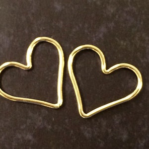 May include: Two gold heart-shaped hoops, one slightly larger than the other, are shown against a dark brown background.
