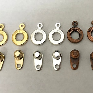 May include: Assortment of 10 metal rings and toggles in gold, silver, and copper finishes. The rings have a circular shape with a small hole in the center. The toggles have a rounded top and a small loop at the bottom. All pieces are marked with "PAT 2007".