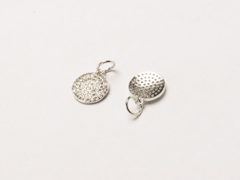 May include: Two silver charms, one with a round, textured surface and the other with a round surface covered in small, clear stones.
