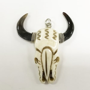 Peut inclure: Un pendentif en forme de cr&acirc;ne de taureau blanc sculpt&eacute; avec des cornes noires et des accents de fil argent&eacute;. Le cr&acirc;ne pr&eacute;sente un design d&eacute;taill&eacute; avec des lignes grav&eacute;es et un petit trou en haut pour l'accrocher.