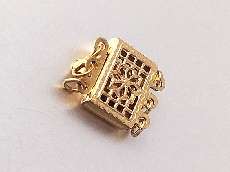 May include: Gold-toned metal clasp with a decorative, openwork design. The clasp has three loops for attaching to a necklace or bracelet.