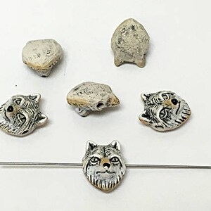 May include: A collection of handmade beads in various shapes and sizes. The beads include wolf faces and abstract shapes, all in a neutral color palette of white, beige, and brown. One bead is strung on a silver-colored pin.