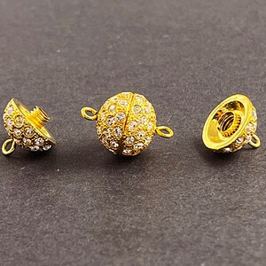 Op de afbeelding: Drie goudkleurige metalen sluitingen met heldere kristallen versieringen. Elke sluiting heeft een schroefontwerp en een lus om aan een ketting of armband te bevestigen.
